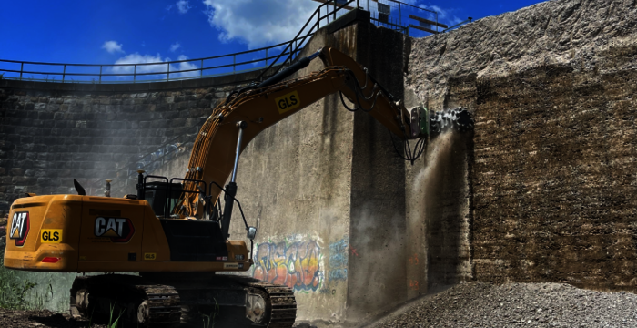Bright yellow CAT excavator with a long hydraulic arm demolishes material inside a circular brick wall, lifting debris as dust rises. A clear blue sky with clouds overlooks the scene.