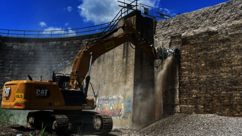 Ein gelber CAT-Bagger arbeitet an einer steinernen Ufermauer und gräbt Erde aus dem Bereich vor der Mauer. Kies und Geröll liegen auf dem Boden; der Himmel ist klar blau mit einigen Wolken.