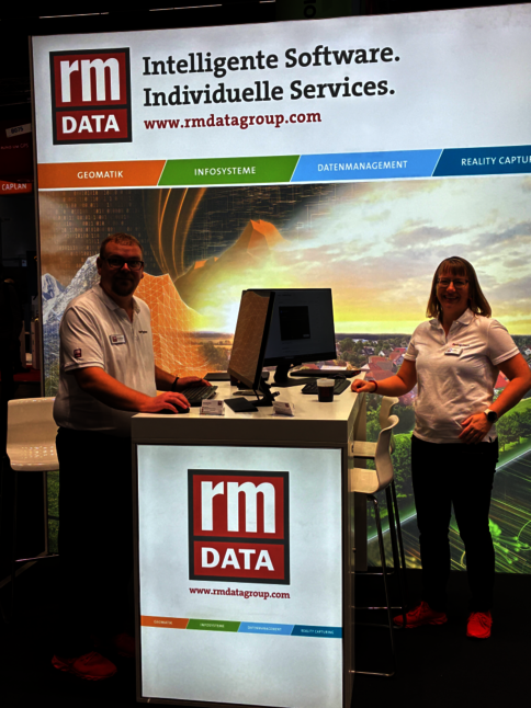 Messestand von rm DATA mit zwei Mitarbeitern (Mann links, Frau rechts). Hinter der Theke ein Computerbildschirm; im Hintergrund Banner mit Logo und dem Satz 'Intelligente Software. Individuelle Services' sowie farbige Kategorienleisten (Geomatik, Infosysteme, Datenmanagement, Reality Capture).