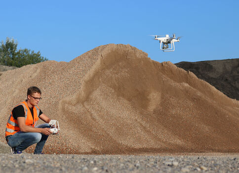 Un homme portant un gilet de sécurité orange est accroupi à côté d'une grande pile de gravier; il tient une télécommande tandis qu'un drone blanc vole près du ciel bleu.