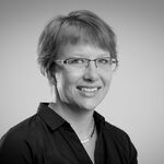 Schwarzweißes Porträt einer lächelnden Frau mit kurzen Haaren, Brille und schwarzer Bluse. Sie blickt leicht zur Seite vor hellem Hintergrund.