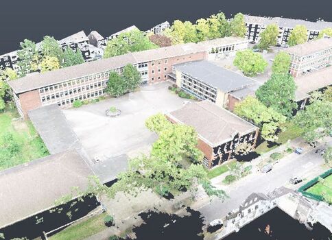 Vue aérienne d'un campus avec plusieurs bâtiments en briques entourant une cour centrale, entourée d'arbres. On aperçoit des espaces verts, des zones piétonnes et des voitures le long des rues, donnant une impression de complexité urbaine.