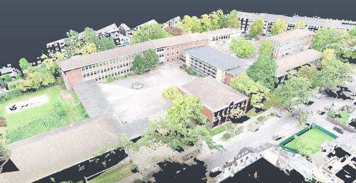 An aerial view of a school campus: several brick buildings around a central open courtyard, surrounded by trees, green lawns, and nearby parking.