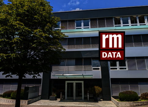 Außenaufnahme eines modernen Bürogebäudes mit Glasfassade. Vor dem Eingang steht eine gläserne Tür; rechts prangt das auffällige rm DATA-Logo an der Fassade, links sieht man einen Baum.