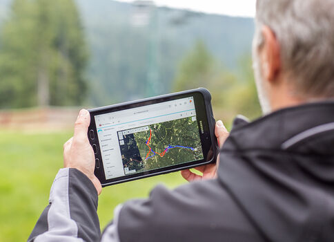 Eine Person von hinten hält ein Tablet, auf dem eine Kartenansicht mit einer markierten Route zu sehen ist; sie steht draußen in einer grünen Landschaft mit Bäumen und Bergen im Hintergrund.