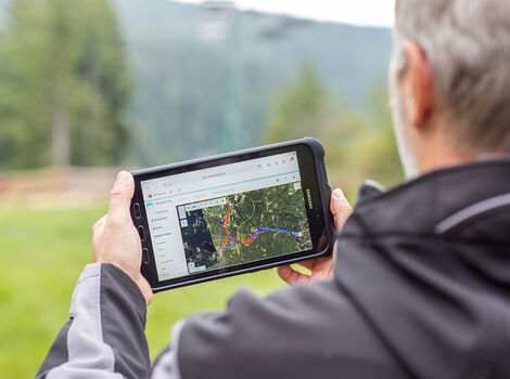 Eine Person von hinten hält ein Tablet, auf dem eine Kartenansicht mit einer markierten Route zu sehen ist; sie steht draußen in einer grünen Landschaft mit Bäumen und Bergen im Hintergrund.