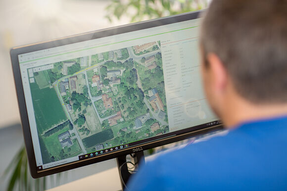 Eine Person sitzt von hinten vor einem Computerbildschirm, der eine Satellitenansicht eines Wohnviertels zeigt – Straßen, Häuser und grüne Flächen.
