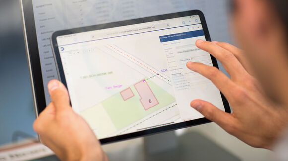 Eine Person hält ein Tablet; darauf ist eine Kartenansicht mit farbigen Feldern, Linien und nummerierten Bereichen zu sehen.