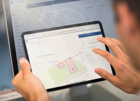 Eine Person hält ein Tablet; darauf ist eine Kartenansicht mit farbigen Feldern, Linien und nummerierten Bereichen zu sehen.