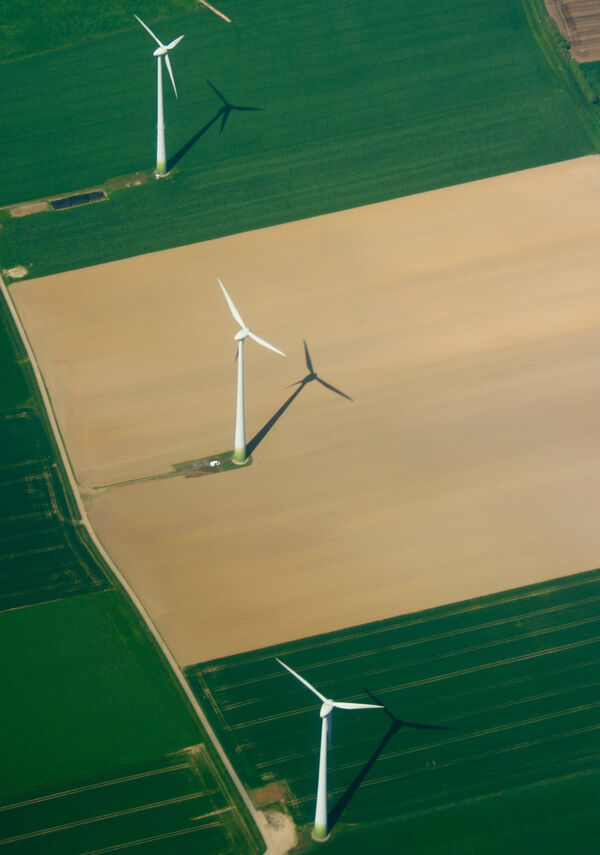 Luftaufnahme von drei weißen Windkraftanlagen auf geteilten Feldern; Rotorblätter ragen hoch und werfen Schatten auf grüne und braune Felder.