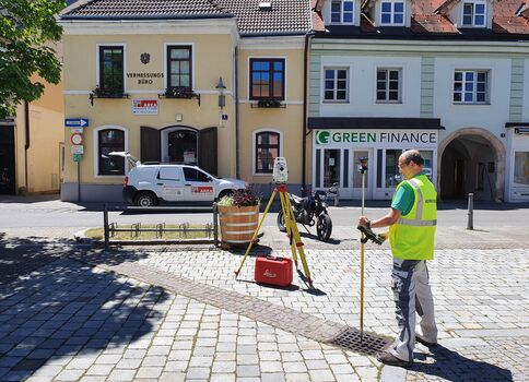 Auf einem Kopfsteinpflasterplatz einer Altstadt steht ein Vermesser in Warnweste und bedient ein Vermessungsgerät auf einem Stativ. Vor ihm liegt eine rote Werkzeugbox und Messausrüstung; im Hintergrund erkennt man gelbe Häuser, ein Vermessungsbüro und ein Green Finance-Geschäft.