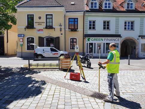 Auf einem Kopfsteinpflasterplatz einer Altstadt steht ein Vermesser in Warnweste und bedient ein Vermessungsgerät auf einem Stativ. Vor ihm liegt eine rote Werkzeugbox und Messausrüstung; im Hintergrund erkennt man gelbe Häuser, ein Vermessungsbüro und ein Green Finance-Geschäft.