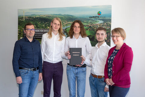Fünf Personen stehen vor einem Banner mit Landschaftsaufnahme; die mittlere Person hält eine schwarze Mappe mit Urkunde, die übrigen lächeln.