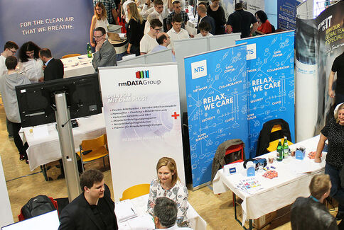 Belebte Messehalle mit mehreren Messeständen, zahlreichen Besuchern, die sich an Infotischen unterhalten. Sichtbare Banner und Firmenlogos kennzeichnen die Stände, während Menschen über Produkte und Dienstleistungen informieren.