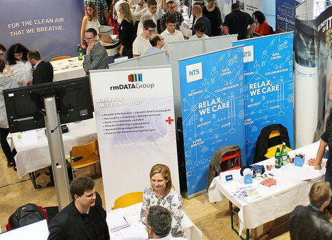 Belebte Messehalle mit mehreren Messeständen, zahlreichen Besuchern, die sich an Infotischen unterhalten. Sichtbare Banner und Firmenlogos kennzeichnen die Stände, während Menschen über Produkte und Dienstleistungen informieren.