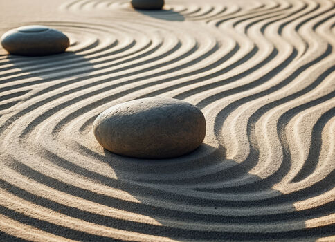 Mehrere glatte Steine ruhen auf feinem Sand in einem Zen-Sandgarten. Der Sand ist kunstvoll in gleichmäßige, wellenförmige Rippen gerissen, wodurch eine ruhige, meditative Landschaft entsteht.