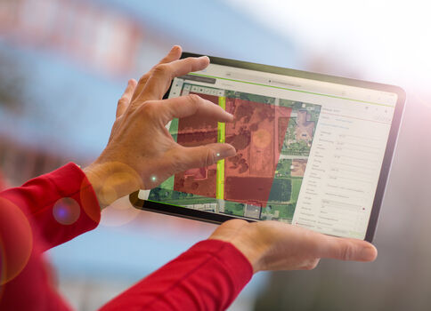 Nahaufnahme einer Person in roter Kleidung, die ein Tablet im Querformat hält und darauf eine Karten-/Satellitenansicht bearbeitet. Die Bildschirmfläche zeigt farblich markierte Felder und ein seitliches Menü.