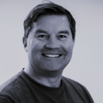 Porträt eines lächelnden Mannes mit kurzen dunklen Haaren, graues T‑Shirt, Blick geradeaus, heller Hintergrund.