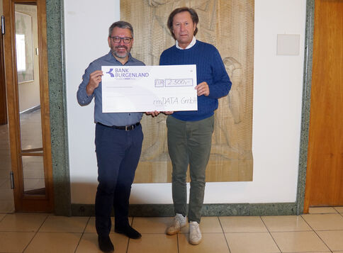 Zwei Männer stehen in einer Eingangshalle und halten gemeinsam einen großen Scheck von der Bank Burgenland; im Hintergrund eine reliefartige Wanddekoration.