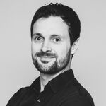 Schwarzweiß-Porträt eines Mannes mit dunklem Haar und Bart, trägt schwarzes Hemd, schaut leicht zur Seite und lächelt freundlich vor hellem Hintergrund.