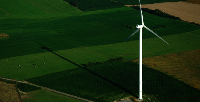 Eine weiße Windkraftanlage ragt über einer patchworkartigen Felderlandschaft; drei Rotorblätter drehen am Turm, der einen langen Schatten über das grüne Ackerland wirft.