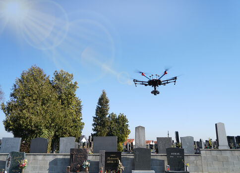 Eine Drohne schwebt über einem Friedhof mit Grabsteinen, im Hintergrund stehen Bäume und ein heller, blauer Himmel mit Sonnenlicht.