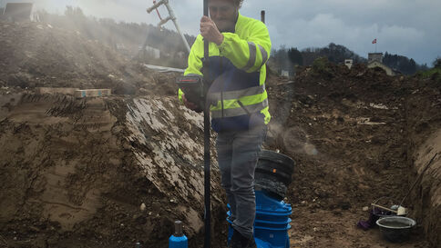 Bauarbeiter in leuchtend gelber Sicherheitskleidung arbeitet in einem tiefen Erdgraben und benutzt ein langes Werkzeug zur Bodenverdichtung; im Hintergrund Erdreich, Staub und Baumaschinen.