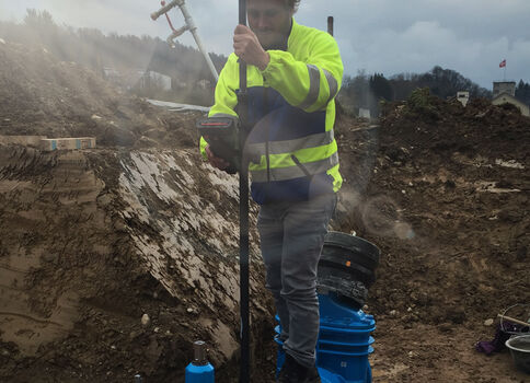 Bauarbeiter in leuchtend gelber Sicherheitskleidung arbeitet in einem tiefen Erdgraben und benutzt ein langes Werkzeug zur Bodenverdichtung; im Hintergrund Erdreich, Staub und Baumaschinen.