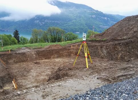 Sur un chantier, une station de relevé sur trépied jaune est installée sur un terrain excavé. À l’arrière-plan, le lac et les montagnes verdoyantes encadrent le site, avec des tas de terre et du gravier.
