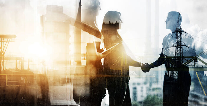 Zwei Geschäftsleute schütteln sich die Hände, vor einer urban-industriellen Skyline im Sonnenlicht.