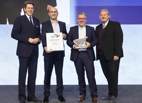 Quatre hommes en costumes se tiennent sur scène lors d’une remise de prix. Deux d’entre eux tiennent un diplôme et un trophée respectivement, tandis que les autres posent et sourient, devant un fond lumineux et un écran.
