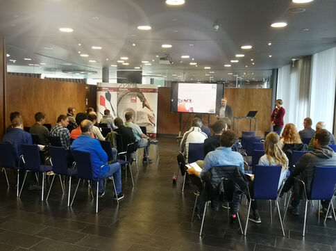 Eine Konferenzraum-Szene: Dutzende Teilnehmende sitzen in mehreren Reihen blauer Stühle und hören einem Redner zu, der am Podium vor zwei Bildschirmen steht und eine Präsentation zeigt.