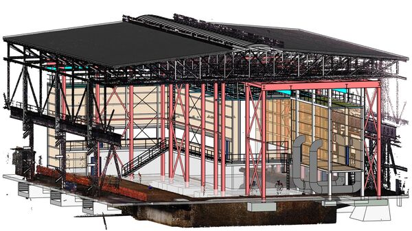 Großes Bauwerk aus Stahlgerüst: mehrstöckige Struktur mit rotem Tragwerk und schwarzem Flachdach. Baustelle mit Betonfundamenten, Baugerüsten und Installationsleitungen im Umfeld, sichtbar sind Rohre und Krananlagen.