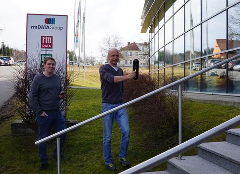 Außenaufnahme: Zwei Männer stehen vor einem modernen Glasgebäude mit Rampe und Treppe. Der rechte Mann hält einen schwarzen Gegenstand in der Hand und blickt zur Kamera; der linke Mann hat die Hände in den Taschen. Ein rmDATAGroup-/rmDATA-Schild ist sichtbar.