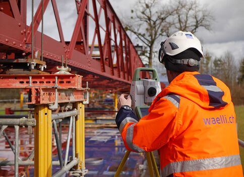 Un ouvrier en veste haute visibilité orange et casque blanc se tient près d'une structure métallique rouge, une charpente de pont, et utilise un instrument de mesure sur trépied, probablement une station totale, sur un chantier avec échafaudage.