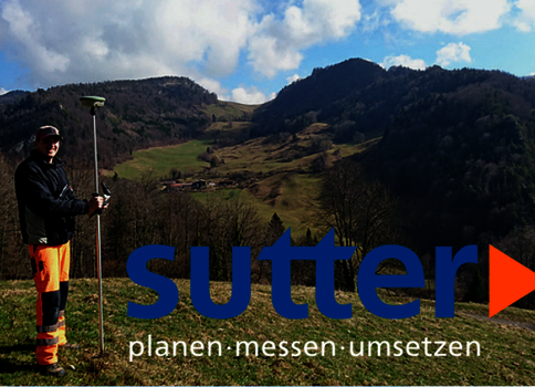 Un homme en tenue de chantier orange et noire se tient sur une colline verdoyante et tient un instrument de mesure fixé sur un mât; en arrière-plan, des montagnes et un ciel bleu avec quelques nuages.