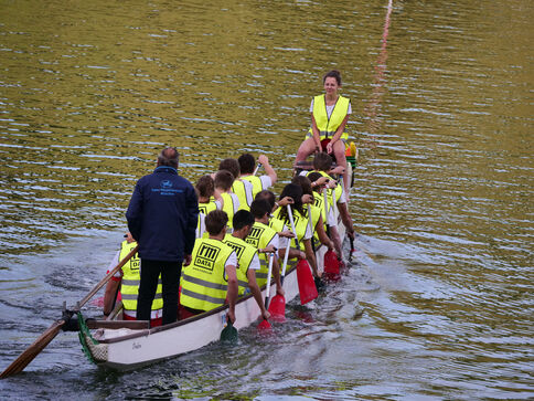 Eine Dragonboot-Crew paddelt synchron in leuchtend gelben Rettungswesten über ruhiges Wasser; das lange, schmale Boot wird von vielen Paddlern getragen.