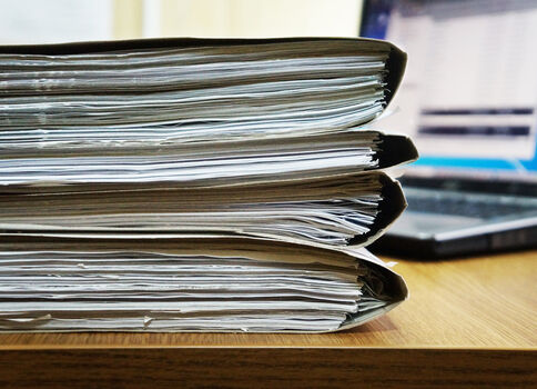 Mehrere Aktenordner mit vielen Papieren stapeln sich auf einem Holztisch; im Hintergrund ist ein Laptop zu erkennen.