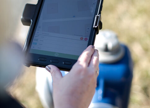 Nahaufnahme einer Person, die ein robustes Tablet in einer Halterung hält; der Bildschirm zeigt eine App mit Textzeilen und Symbolzeilen; Hintergrund unscharf, wirkt im Freien.