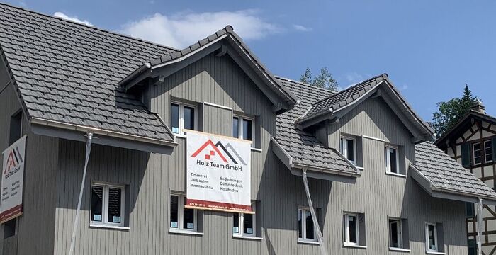 Eine Häuserzeile mit grauen Holzfassaden und Satteldächern sowie mehreren Dachgauben; an der linken Gebäudeseite hängt ein Werbebanner der Holz Team GmbH.