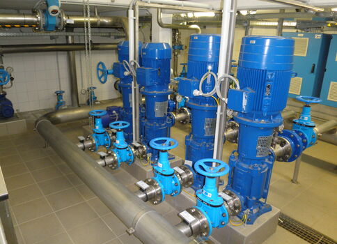 Industriepumpenanlage: Mehrere blaue Pumpenmotoren mit Ventilen und Rohrleitungen in einem Technikraum; sichtbare Handrääder, Steuereinheiten und Verdrahtung.