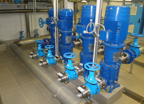 Industriepumpenanlage: Mehrere blaue Pumpenmotoren mit Ventilen und Rohrleitungen in einem Technikraum; sichtbare Handrääder, Steuereinheiten und Verdrahtung.