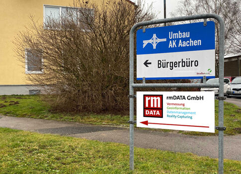Auf einem Gehweg steht ein Metallschild mit blauem Hinweis „Umbau AK Aachen“ und Pfeil nach links zum Bürgerbüro.