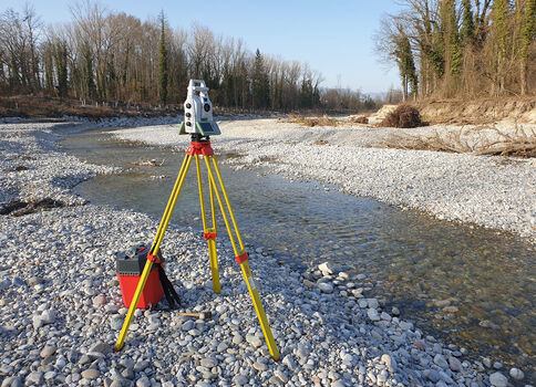 Trépied jaune de relevé installé sur une berge rocheuse près d'une rivière peu profonde; un instrument de mesure métallique est fixé dessus et une mallette rouge est posée à côté dans un paysage forestier.