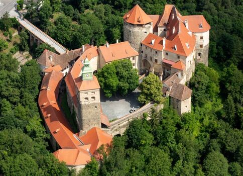 Luftaufnahme einer mittelalterlichen Burganlage in dichtem Wald: mehrere Türme mit roten Ziegeldächern, befestigte Mauern um einen Innenhof; die Anlage wirkt wie eine Festung mitten im Grün.