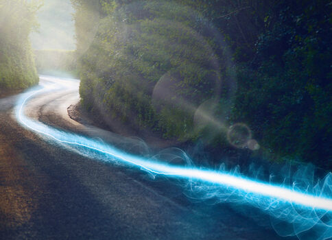 Eine kurvige Straße windet sich durch einen dichten Wald. Sonnenlicht fällt durch die Bäume, während eine leuchtend blaue Lichtspur die Straßenseite entlang zieht.