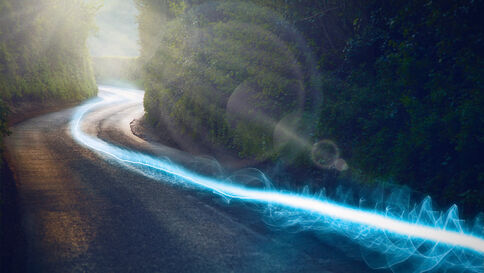 Eine kurvige Straße windet sich durch einen dichten Wald. Sonnenlicht fällt durch die Bäume, während eine leuchtend blaue Lichtspur die Straßenseite entlang zieht.