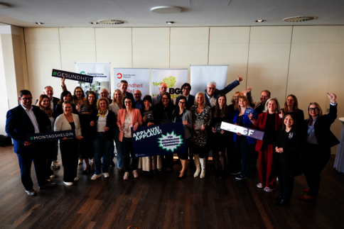 Eine gemischte Gruppe von Menschen posiert in einem hellen Raum, hält Schildchen und Banner mit Gesundheitsbotschaften; im Hintergrund stehen weitere Banner. Die Stimmung ist fröhlich und alle schauen in die Kamera.