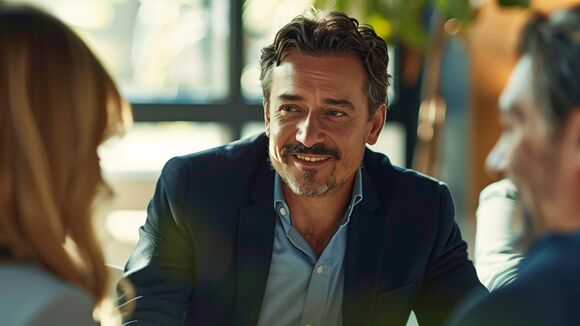 Ein gut gekleideter Mann mit Bart lächelt während eines Gesprächs mit zwei Personen in einem hellen, modernen Raum.