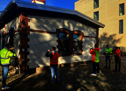 Kleines Haus mit weißer Fassade, dunklem Dach und roten Backstein-Akzenten; mehrere Personen in leuchtend gelben Sicherheitswesten fotografieren das Gebäude im Außenbereich.